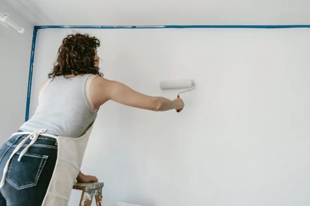 Frau streicht auf einer Leiter eine Wand mit der Farbrolle weiß, die Kanten sind abgeklebt.